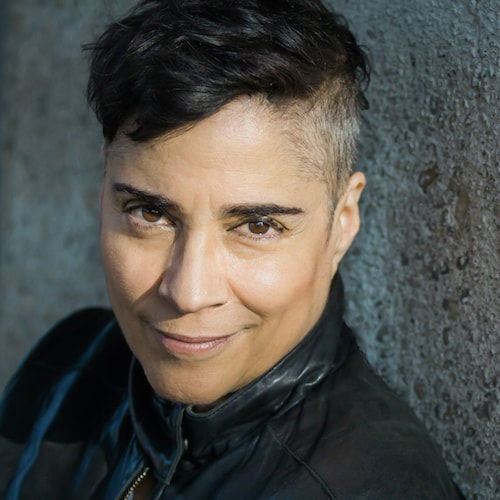 A headshot of a woman with olive skin and short, dark hair that is cropped close at the sides. She is looking up at the viewer with a small smile. She wears a black leather jacket and stands in front of a grey wall.