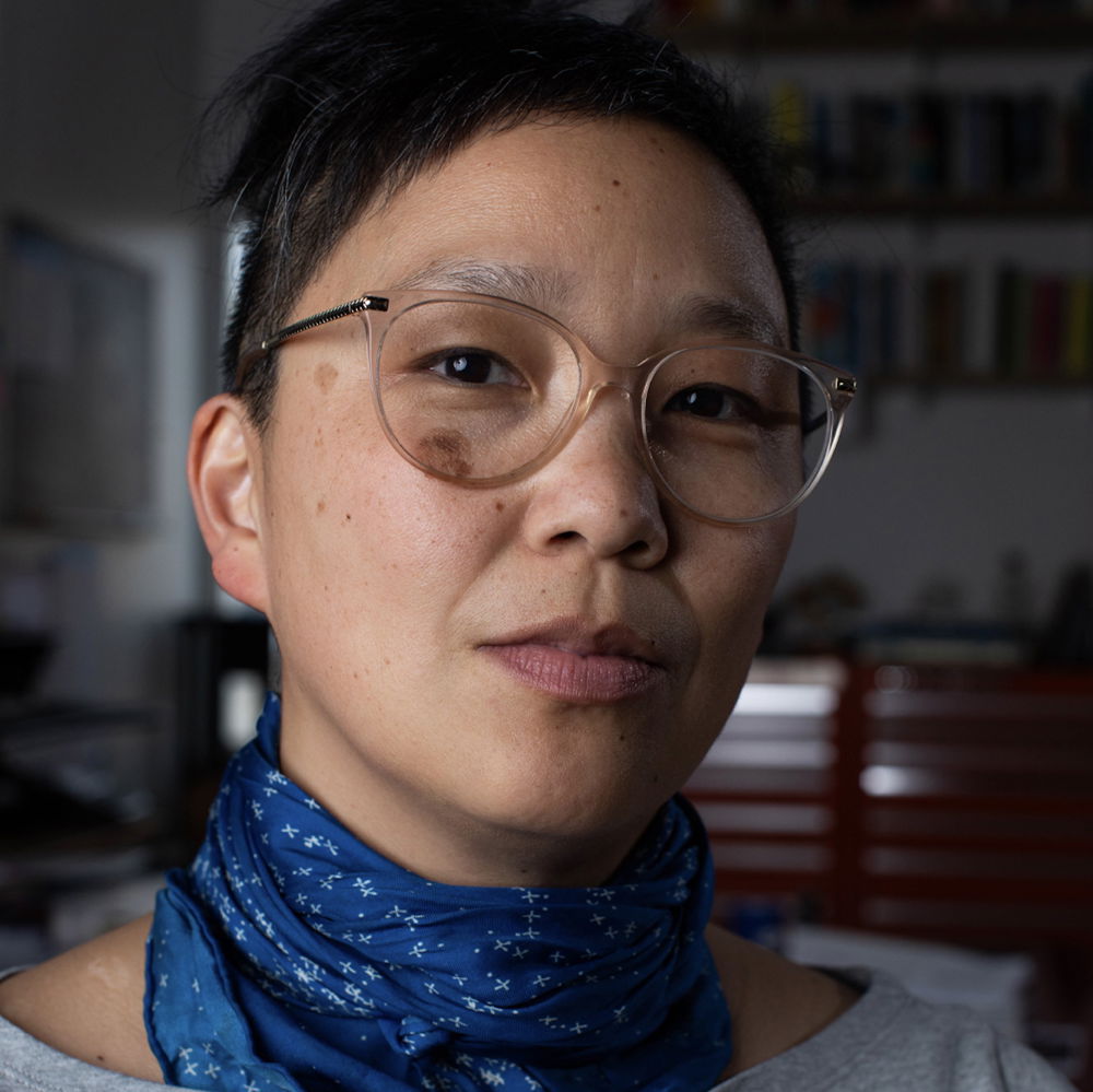 An Asian woman with transparent-frame glasses, blue scarf, and light gray shirt sitting for portrait.