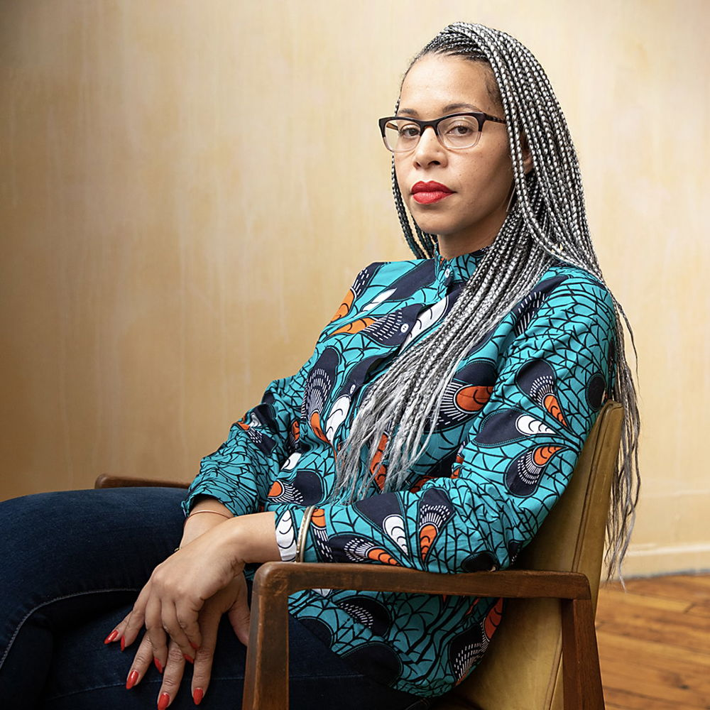 A Black woman with a light brown complexion wears glasses and a turquoise blouse with dark-blue and orange accents. Her long hair is in silver braids that reach to her mid-back. She sits in front of a yellow background looking decisively at the camera.
