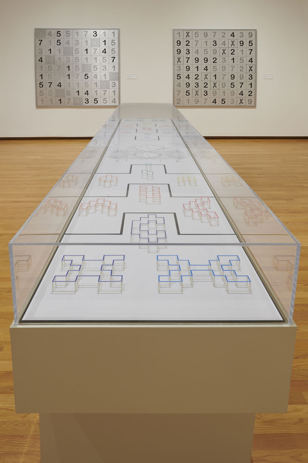 A long rectangular display case with intimate-sized plexiglass sculptures laid flat on their white pedestal. The geometric abstract shapes resemble pixelated letters found in the English Alphabet. The top of the plexiglass is outlined in different bright colors.