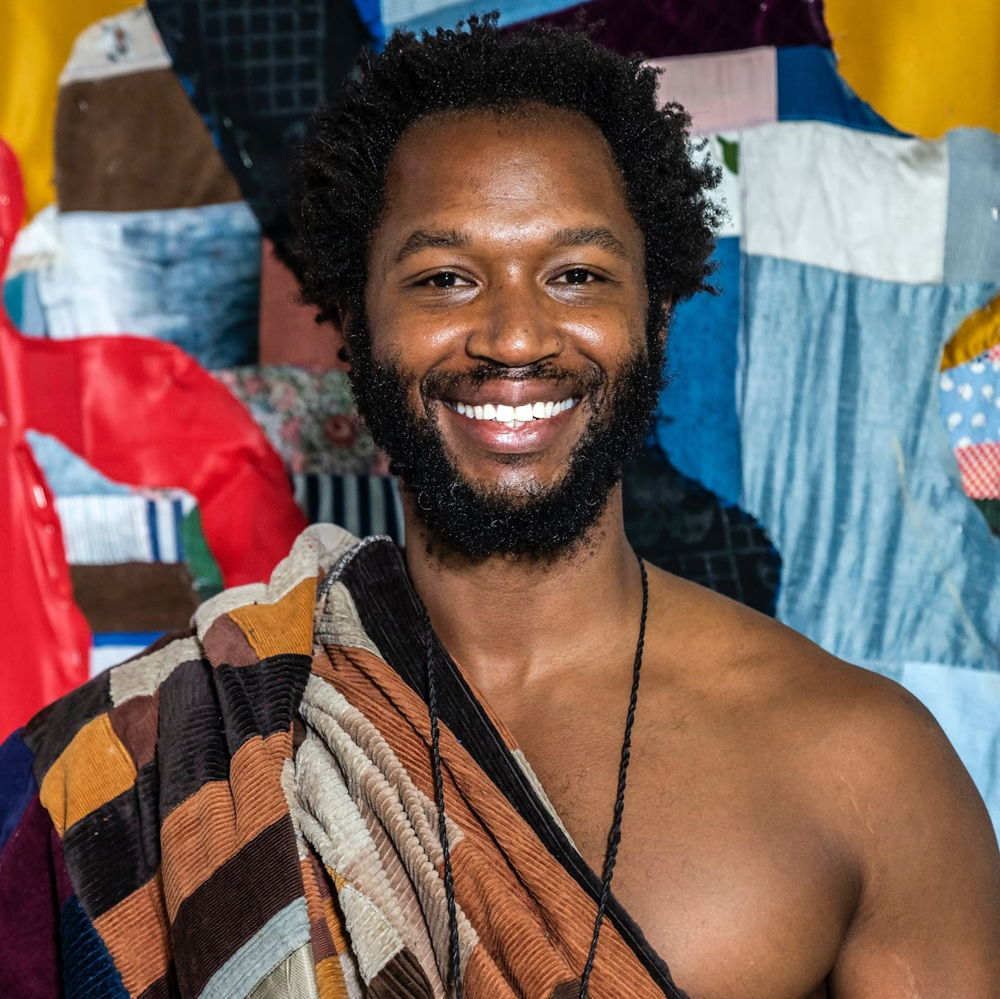 Basil, an African American, is pictured from the waist up, smiling, standing in front of one of Basil’s quilt works, which hangs on the wall behind the artist. The hanging work is a myriad of colors. Basil is also wearing a quilt, another one of Basil’s works, which is draped over Basil’s body exposing one of Basil’s shoulders.