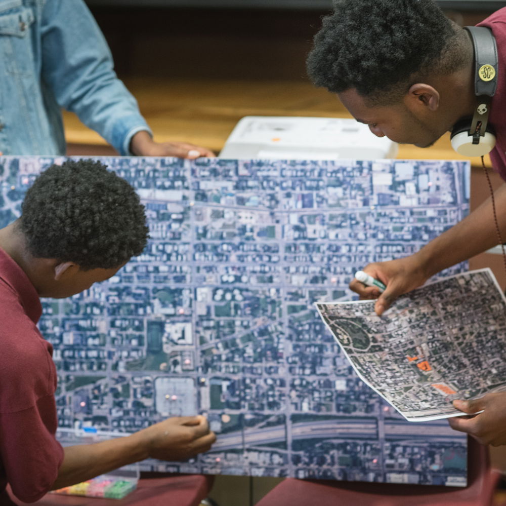 Two Black teens in school uniforms study a bird's eye view map together. The map shows  several blocks and buildings as well as a highway.