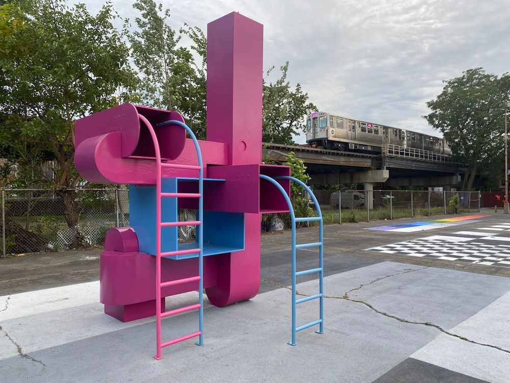 Photo of a pink and blue jungle gym installation with tubes resembling an HVAC system and two ladders. Behind the structure, an elevated train is going by.