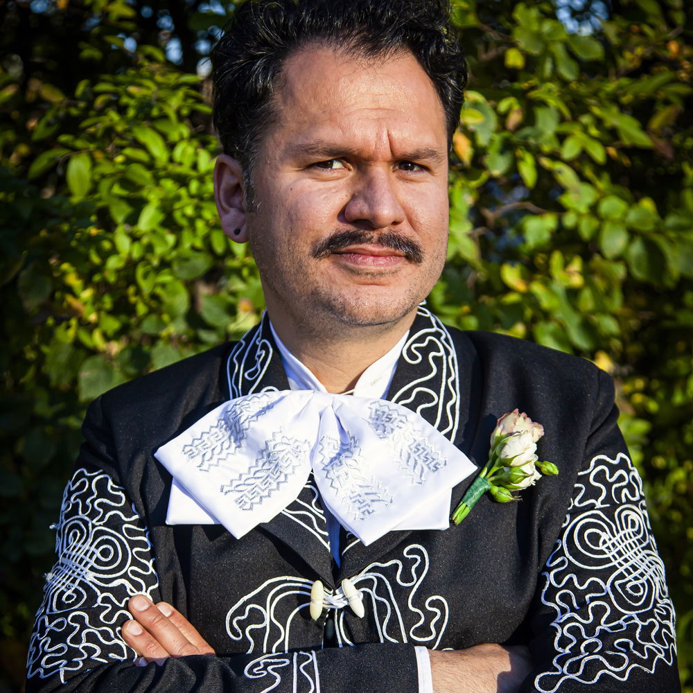 Salvador, a Latinx man with dark wavy hair and a black mustache, stands against a green bush. He is wearing a mariachi suit without the hat.