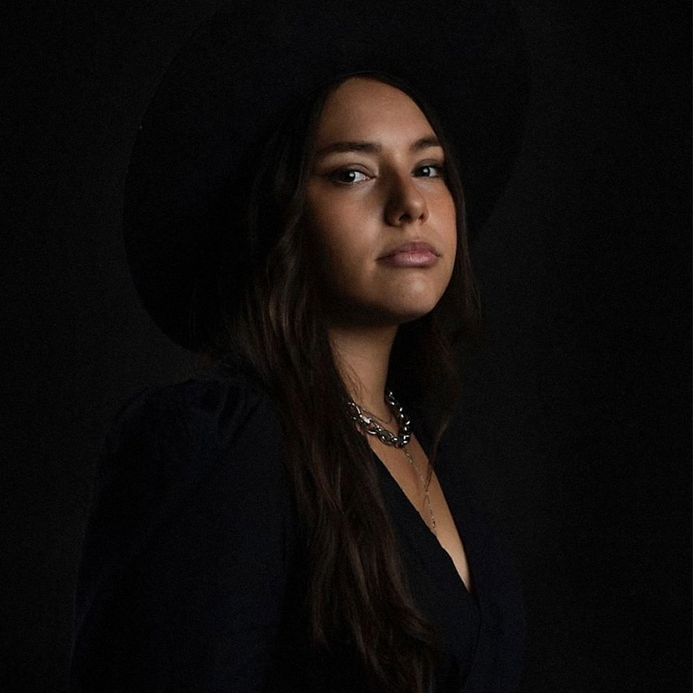 Headshot of a woman with tan skin and long brown hair in front of a dark background. She wears a black long-sleeved dress, silver chain, and wide-brimmed black hat.