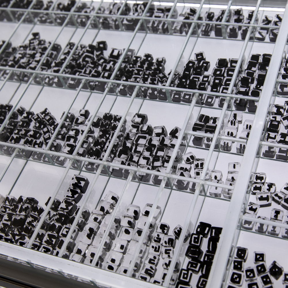 A black-and-white photograph of small glass cubes organized in clear acrylic containers. The small cubes have subtle differences by which they are organized; ranging from completely clear to clear with the addition of black lines.