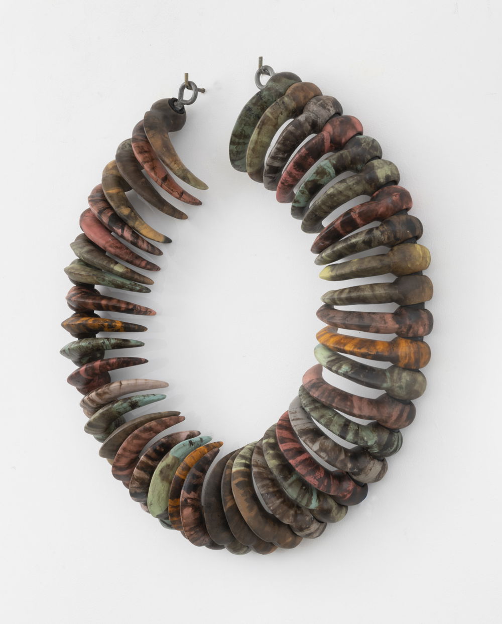 A wreath of curved talons made of polished stones hangs on a white wall. Each talon is uniform in size and shape but is uniquely colored in greens, browns, oranges, and yellows.