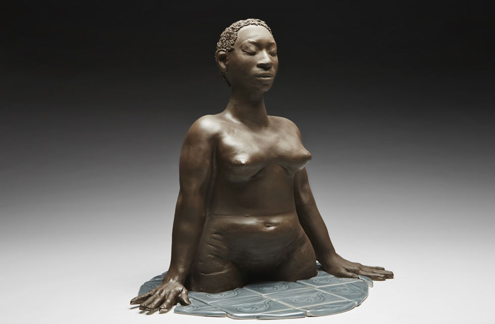 A sculpture of a nude woman from the waist up, her hands resting upon the gray-blue tiles that surround her, as though she is emerging from the water.