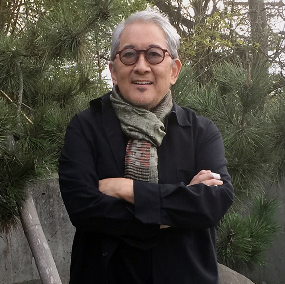 A man with short gray hair and glasses smiles slightly with his arms crossed in front of him. He wears a multi-colored, patterned scarf and a dark pinstriped blazer.