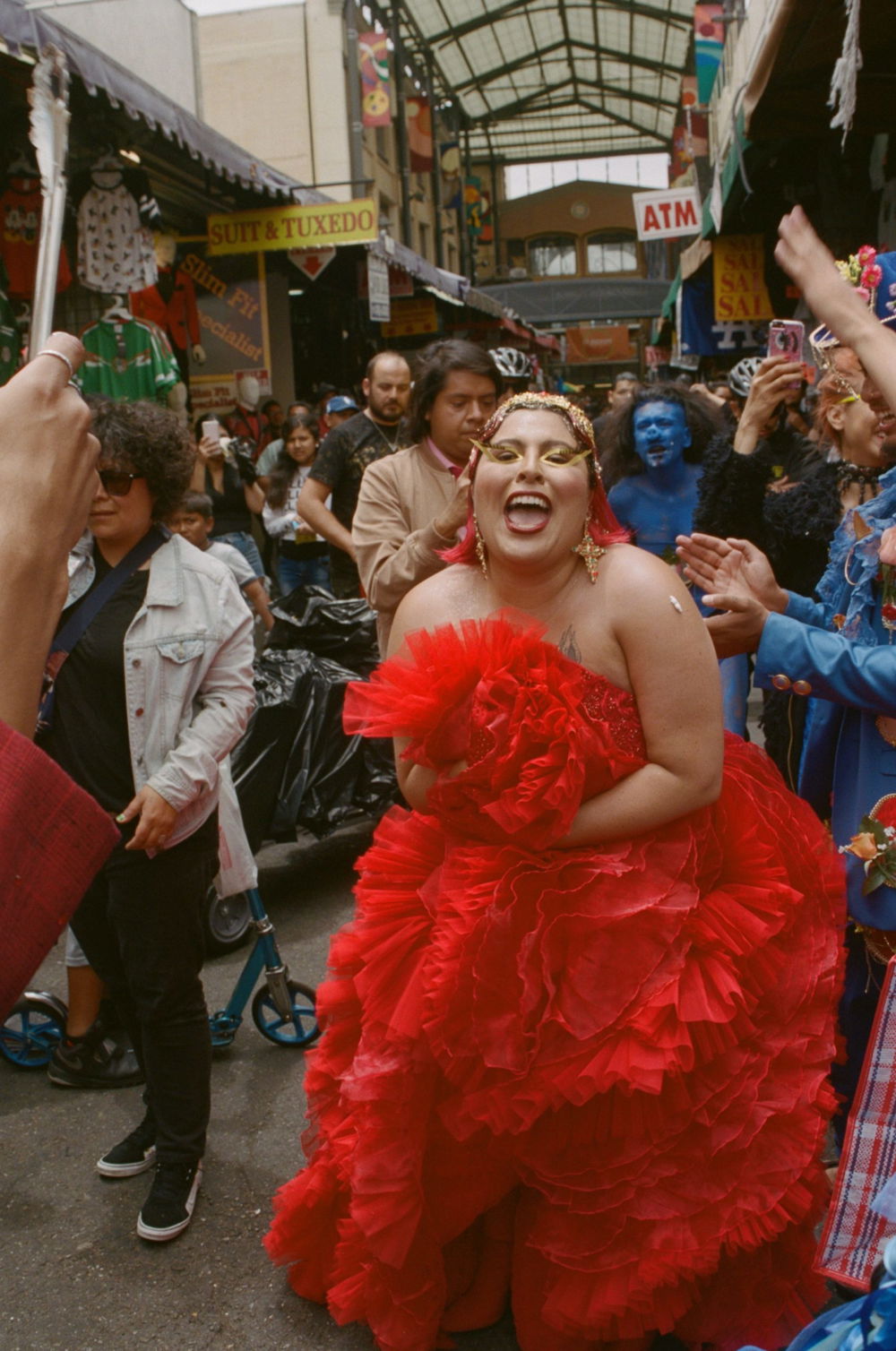 A woman in a bright red dress and matching lipstick smiles widely, surrounded by other people in a covered pedestrian boulevard.