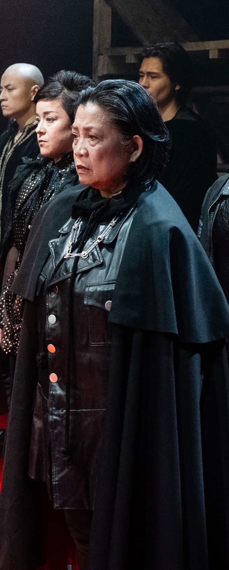 Mia Katigbak, an Asian American woman with combed back shoulder-length black hair, stands on stage and gazes forward with other actors. She wears a collared black leather top under a dramatic black cape.