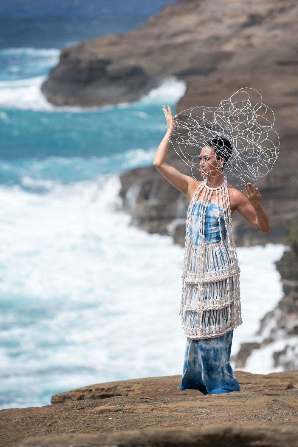 Photo of a woman standing on rocks by the waterfront wearing a white-and-blue foot-length dress. A white woven garment hangs from her neck and descends just past her knees, showing the dyed fabric underneath. Delicate loops of wire form a cloud around her head which she holds up with her hands.