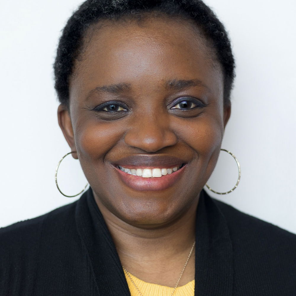 A headshot of a Nigerian-American woman taken against a white background. She has deep brown skin, a coily, close-cropped salt-and-pepper afro, and is smiling directly at the camera. She is wearing a soft black cardigan over a yellow knit sweater, a pair of thin gold hoop earrings, and a delicate gold necklace.