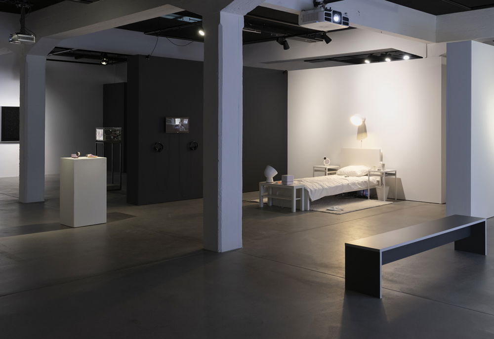 View of an installation in gallery with bed, nightstands, carpet, lights, all in white. On the left is a white pedestal with small purple sculptures set on top of it.