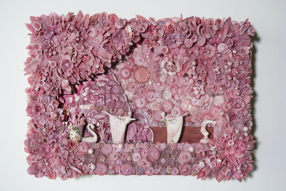 A rectangular-shaped wall sculpture made of all pink glass, resembling a ballet stage with curtains. Two pink ballerinas stand in front of a sunset, ocean, and mountain. On their right is a fish and on the left is a small pitcher painted with flowers.