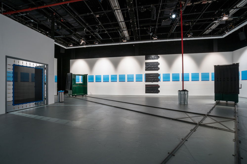 In a gallery space, a dumpster is split in two and each half moves along train rails. The main entrance door is spanned by a trampoline, and trash bags are held up on walls by static electricity.