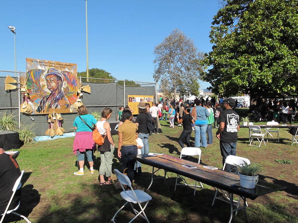 A crowd of people facing away from the camera look at art and posters that have been hung up on wire fencing in a grassy park. Closer to the camera are white plastic chairs around long wooden tables that each have a potted plant on top.