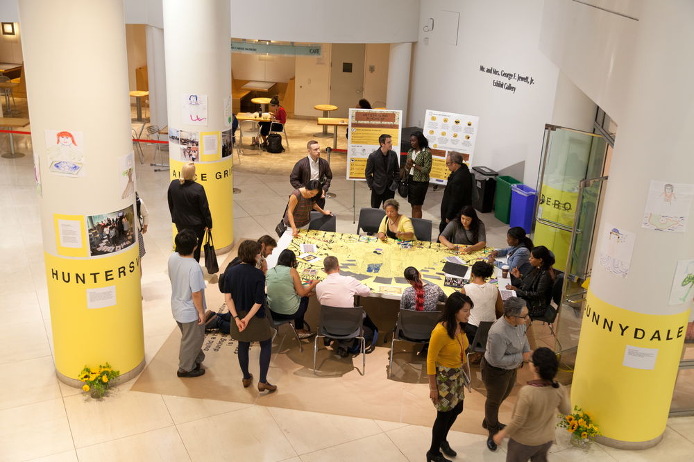 A group of people in business casual attire sit and stand around a table in a large room. Some are drawing on a yellow sheet covering the table while others are standing around the table and talking with each other. There are posters, drawings, and photos displayed around the table.