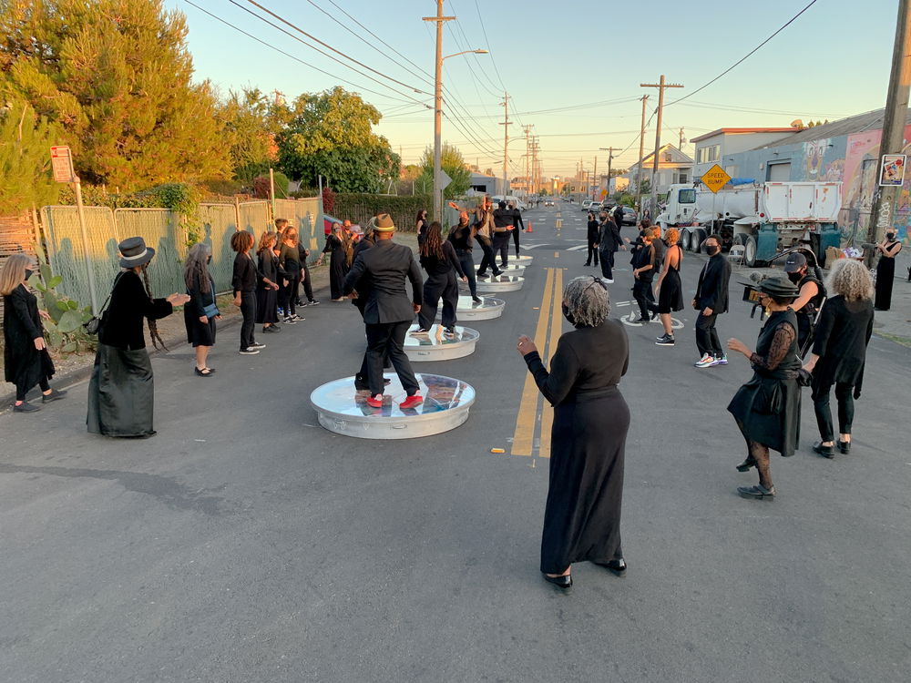 A large group of people in black clothing are gathered in the middle of a residential street during sunset. In the middle of the group are people dancing on raised circular platforms.