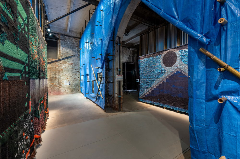 A large installation shot of a long, narrow hallway in an industrial interior. On the left hangs a textile composed of abstract shapes and patterns of black, green, red, and gray. The wall on the right is covered with a blue tarp and protruding bamboo sticks. On this wall an arched entryway leads into a dim gallery space where a large tapestry hangs, depicting a dark-blue pyramid and floating black orb on a background of blues and grays.