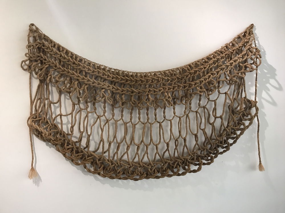 A skirt tightly woven at the top and bottom, with looser loops resembling netting in the middle. It hangs from a white wall, with the ties dangling down each side.