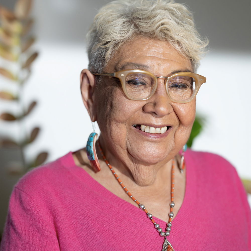 An older Native woman with short gray hair and glasses smiles widely. She wears a pink shirt, a beaded necklace, and earrings.