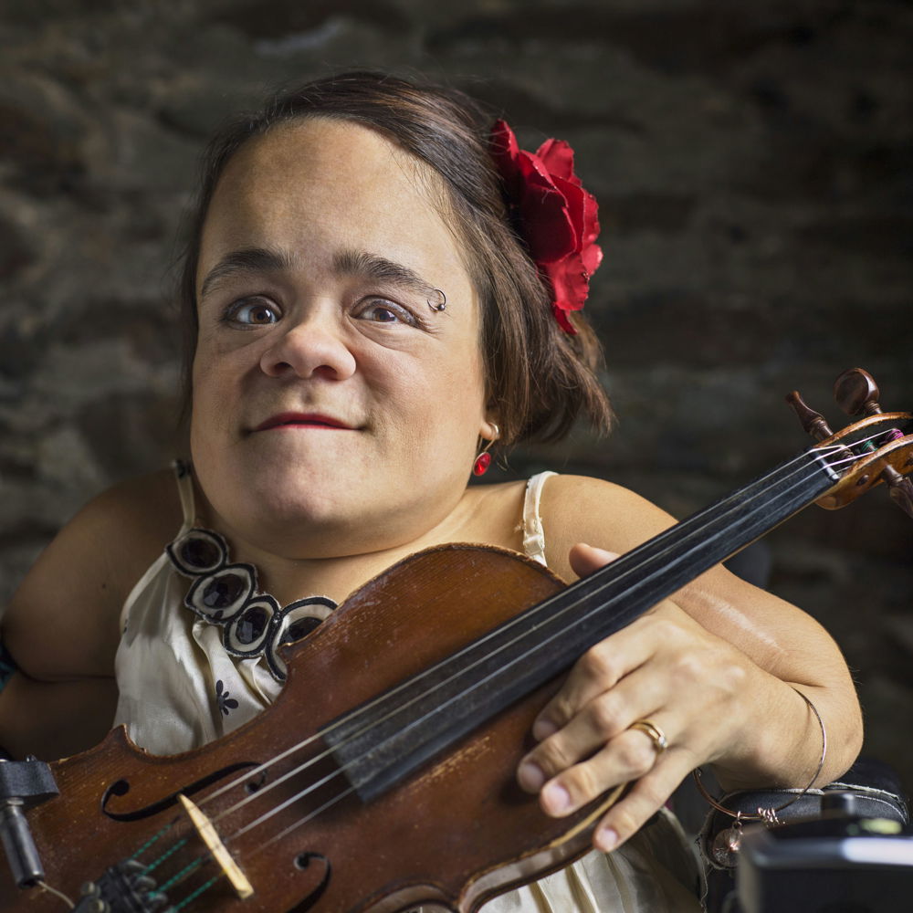 Gaelynn, a small white woman seated in an electric wheelchair, holds her violin and smiles. She has medium-length brown hair held back with a red flower clip and wears a white silky dress with large black beads sewn along the neck.