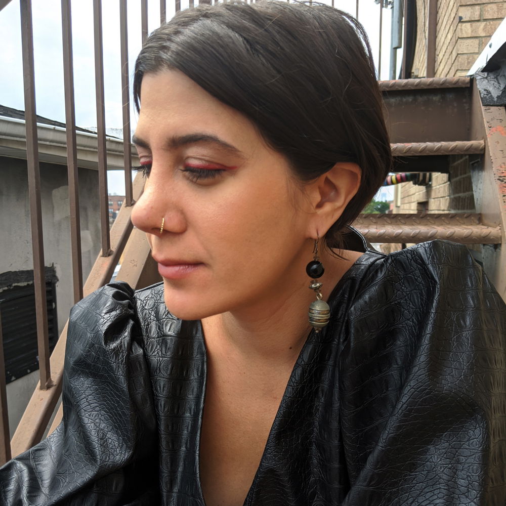 Morehshin, a young Iranian woman with straight dark hair, sits on a staircase. She is wearing a black leather dress with silver and black earrings.