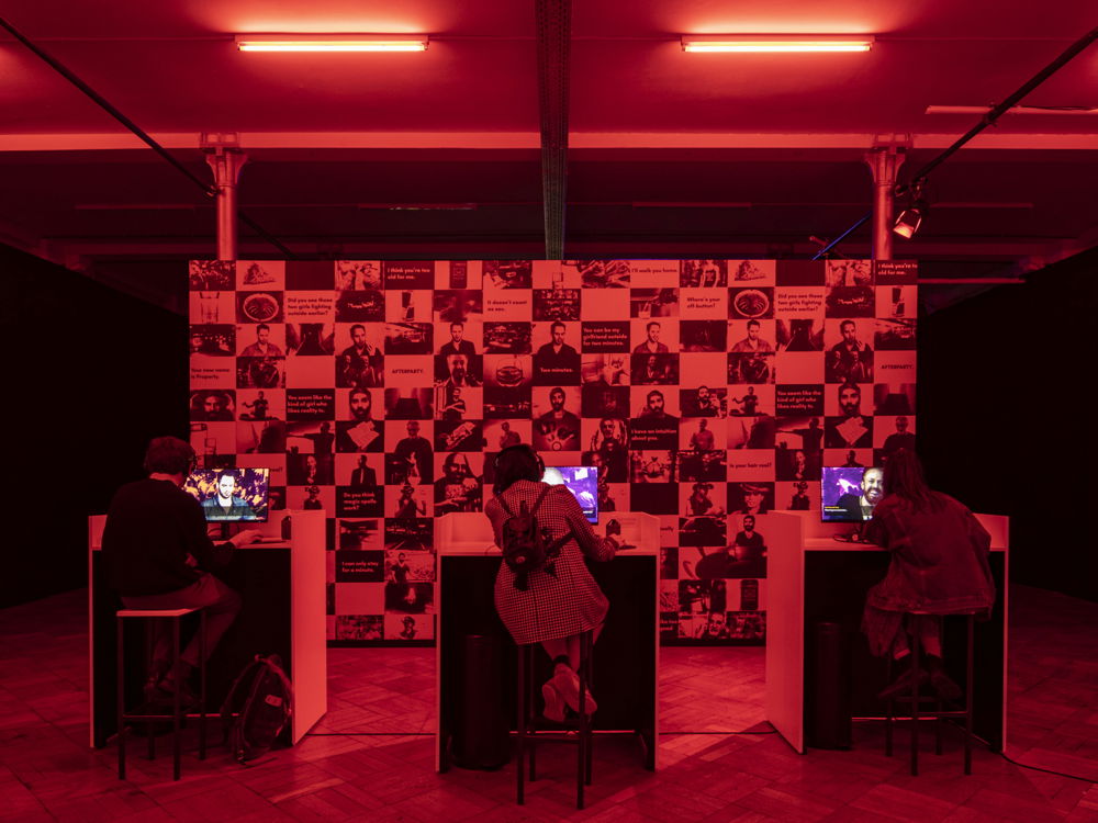 In a​​ windowless room bathed in red light, three people sit with their backs to the camera and facing a computer monitor. In the background is a floating wall covered in tiled images.