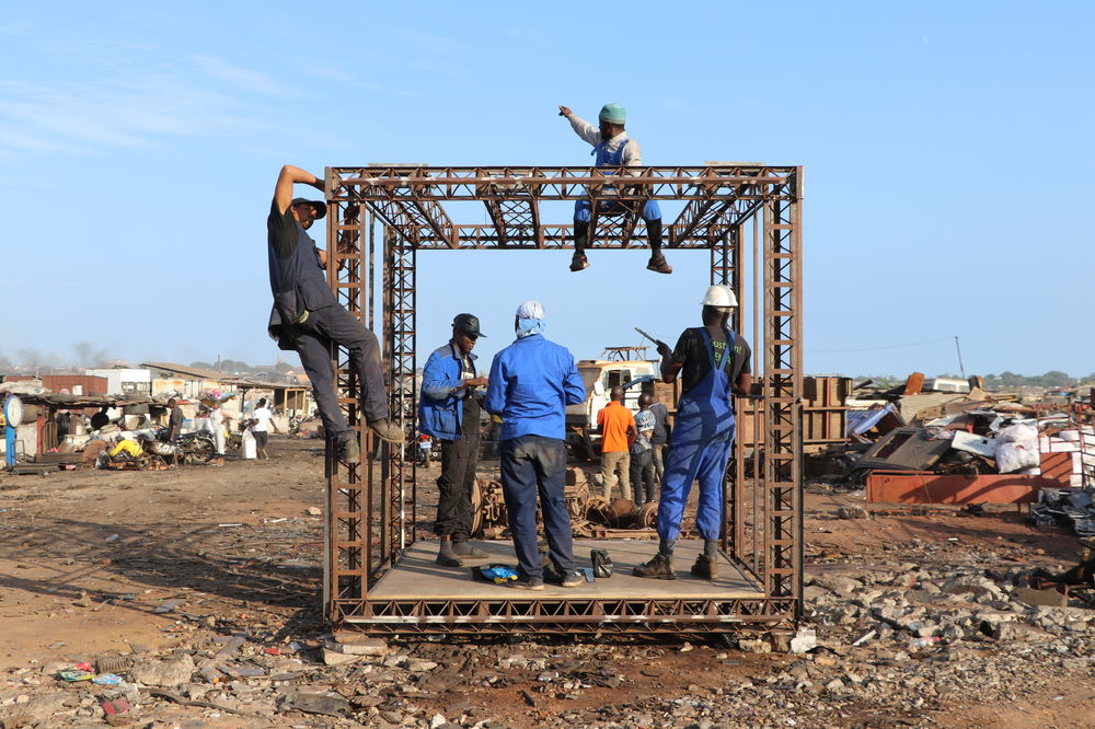 Five men climb inside and on top of a cubic structure. The structure has a small wooden deck underneath lightweight steel webbing and is situated in a scrapyard surrounded by rubble.