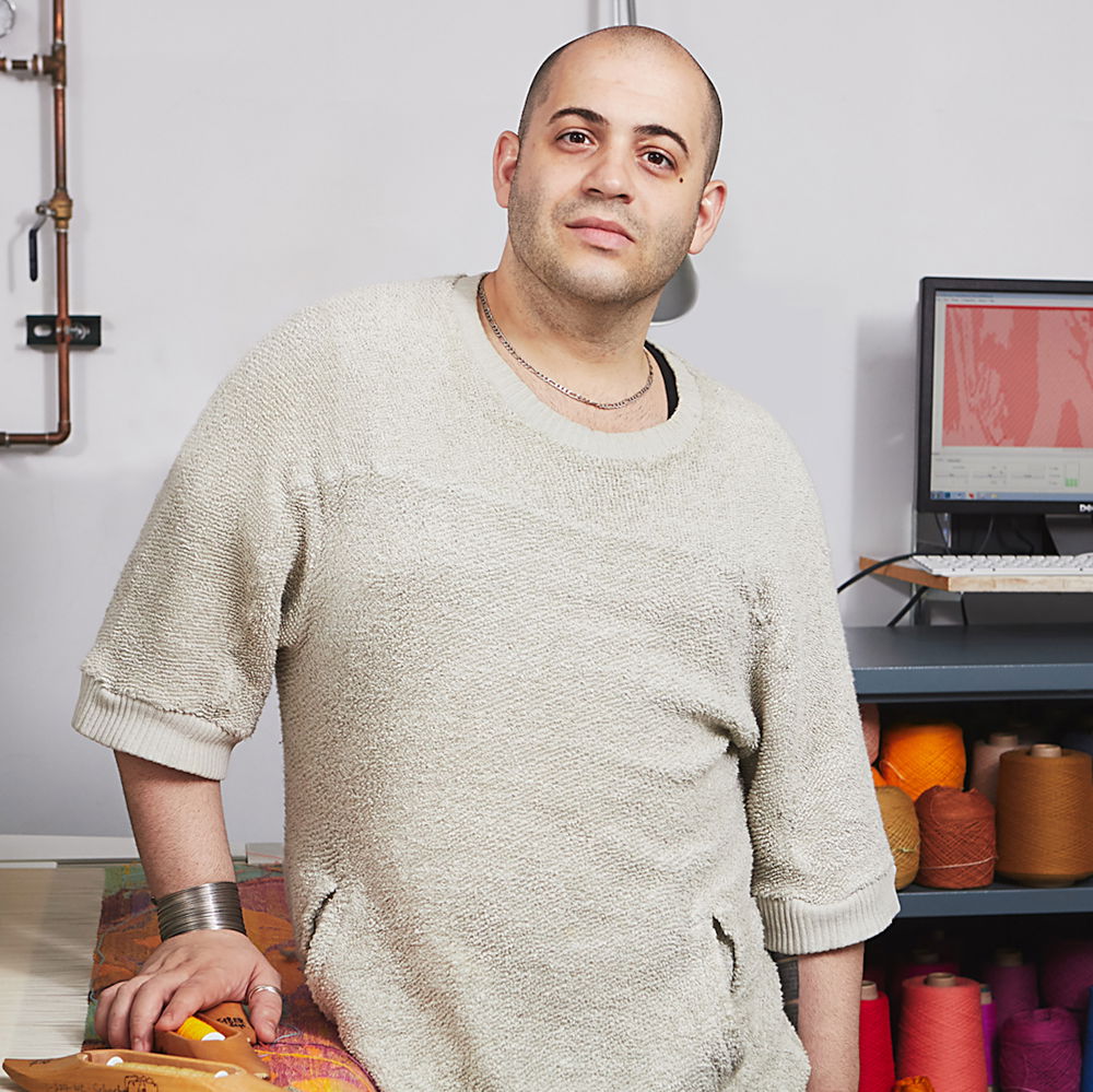 A person in cream-colored sweater leans on a table and gazing into the camera. They are standing in a workspace in front of shelves lined with colorful skeins of thread.