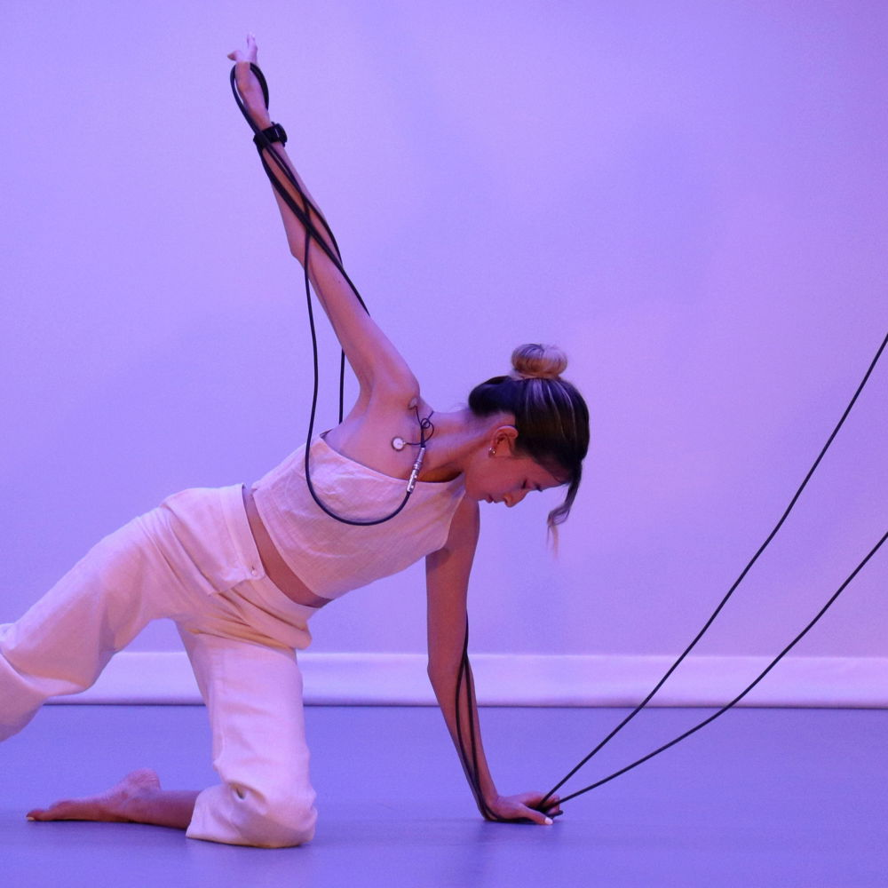Mid-dance, long dark cables extend out of Yo-Yo's body connected to her skin with golden discs. She steadies herself with one hand on the floor and another raised high, her thin arm wrapped in cables, one knee on the floor her other leg extended. She gazes downwards, listening inwards.