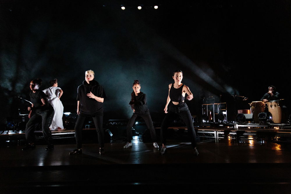 Four people wearing black dance in unison on a dark stage with dramatic lighting. Photographed in mid-motion, they stand with their legs spread apart and their left arms slightly extended.