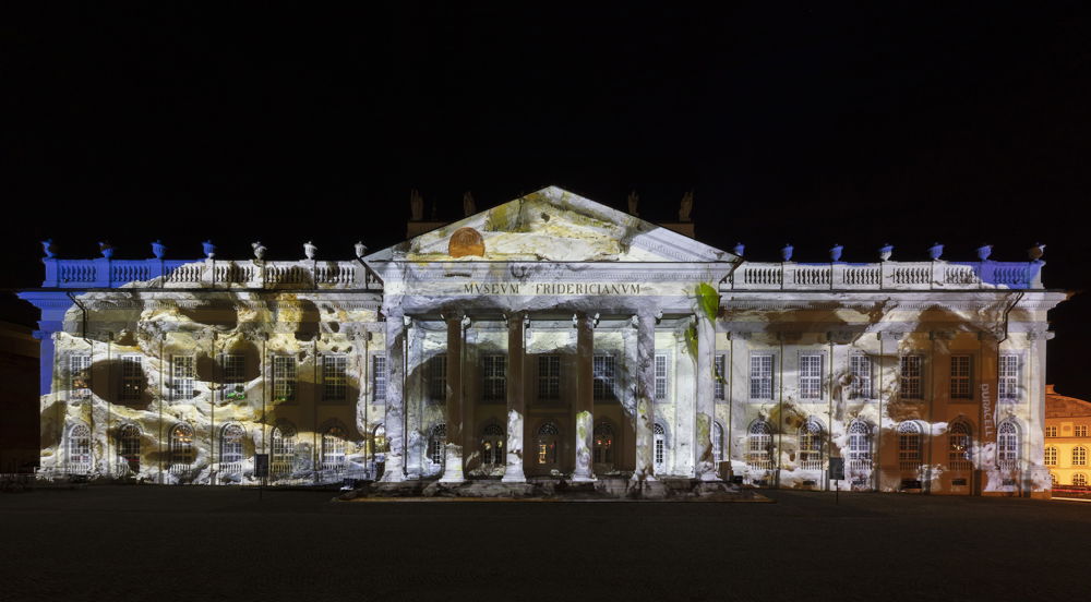 A lit-up museum facade at night time. The museum is neoclassical, imitating a Greek temple. Projected on the museum is an image of a porous sand-colored stone.