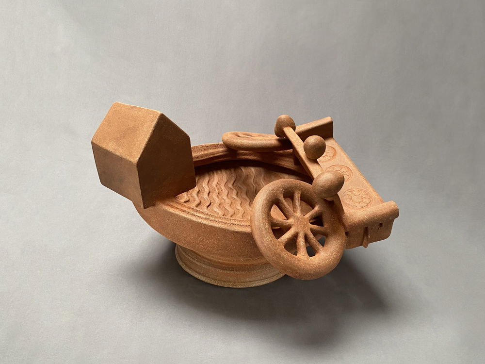 An unglazed red-brown ceramic sculpture of a toppled wagon and house perched around the edge of a bowl-shaped structure.