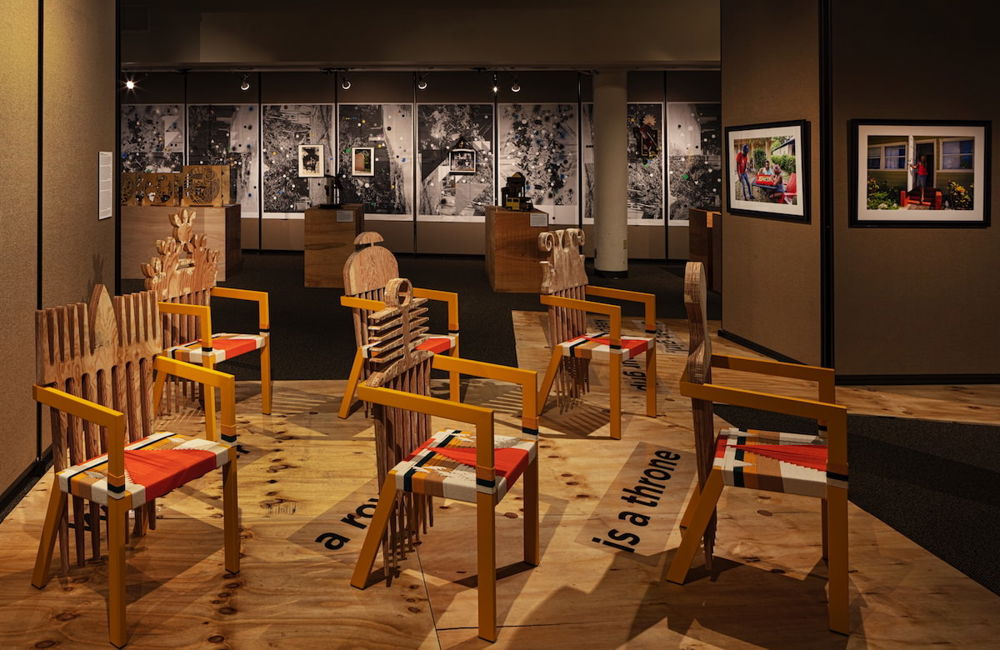 Photo of six chairs set up in a museum space. The backs of the chairs are carved to resemble hair combs, each one in a slightly different style. The seats of the chairs are woven with a red-and-white design.