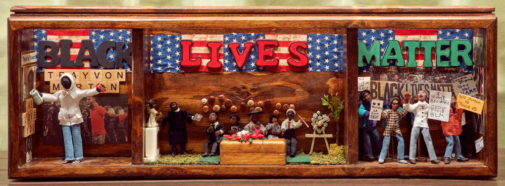 A “trilogy box” of three dioramas. The left diorama depicts Trayvon Martin wearing a hoodie and carrying ice tea and Skittles; behind him are images of Black men that have been killed by police and a protest march. The center diorama depicts a funeral with a Black family burying their child. The mother holds a picture of her daughter. The right diorama depicts a street protest with signs that say “I can’t breathe — George Floyd,” “Sandra Bland — Say Her Name,” and “No Justice No Peace."