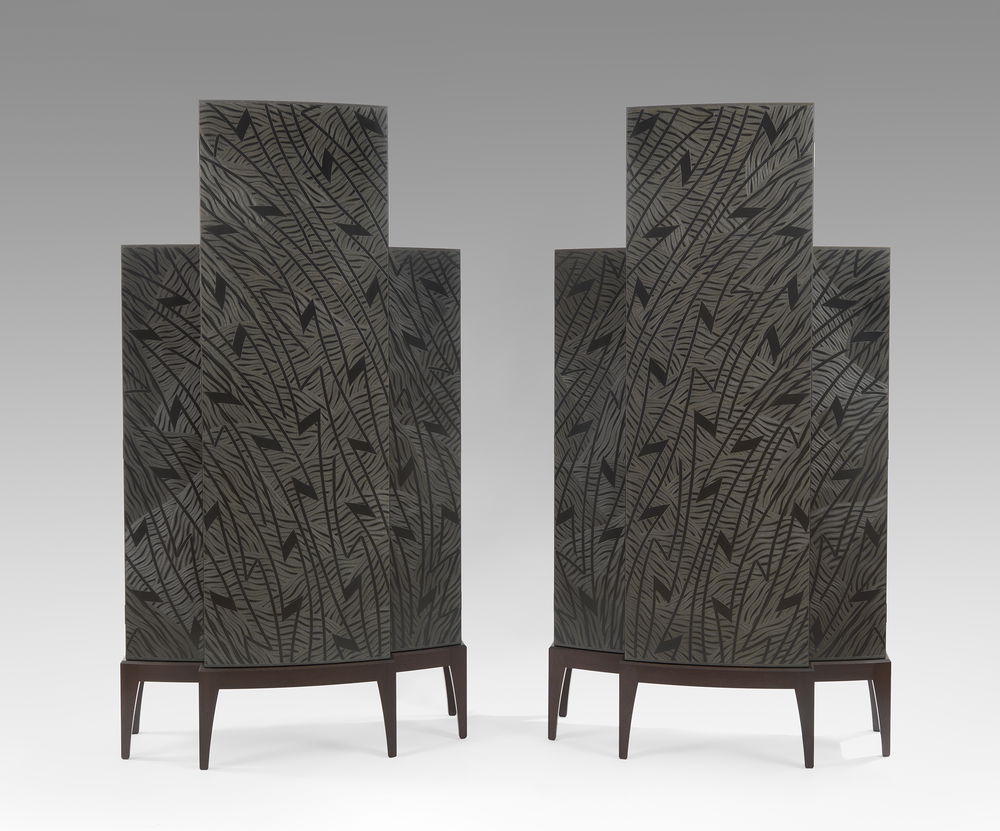 Two patterned black cabinets stand next to each other. They are mirror images of each other, with white zigzag and wavy designs decorating their surfaces. Both are composed of three curved vertical doors, with the tallest door in the middle flanked by two doors that are smaller and equal in width and height. Each cabinet rests on a six-legged stand.