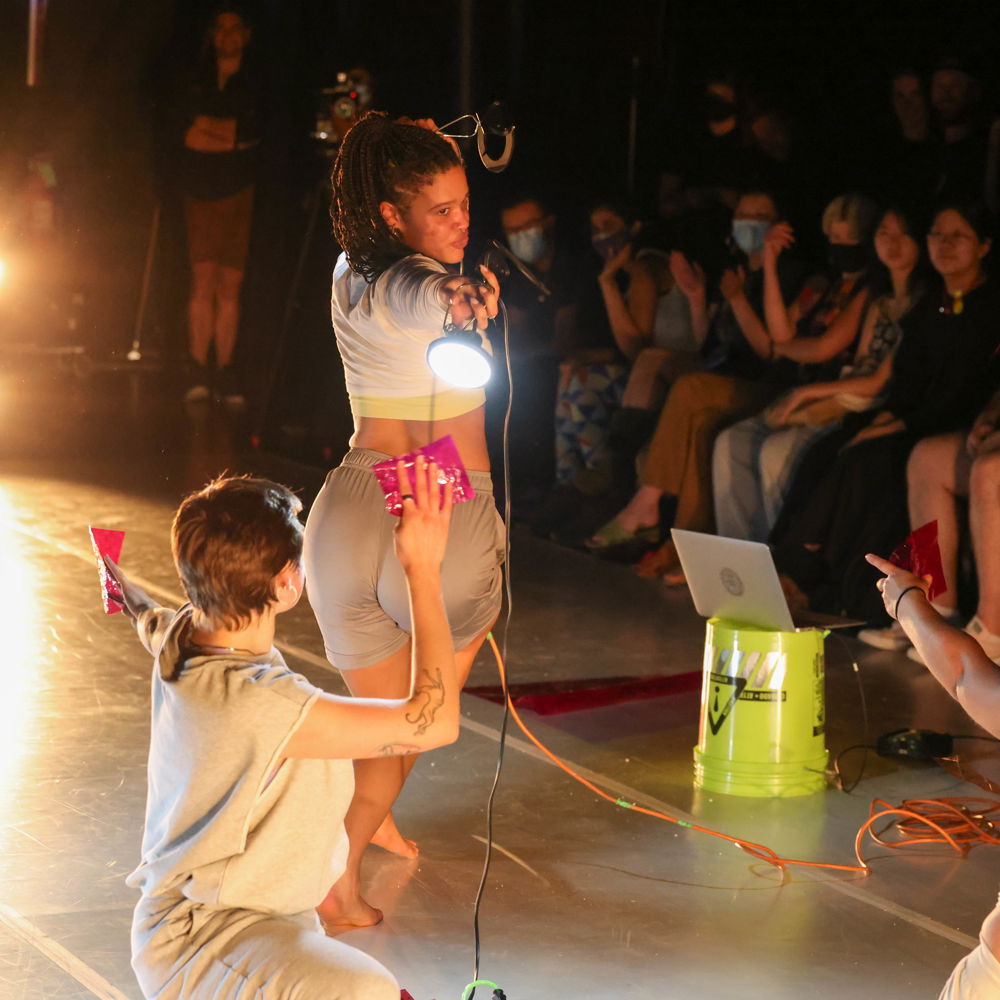 [ID: Still of four performers mid-motion wearing white or gray tees and sweatpants. One of them holds up a work light while the other three toss color gels and filters into the air. More color gels are scattered on the stage floor along with two different colored extension cords and a laptop resting upon a fluorescent bucket. Behind them, the first audience row lines the length of the stage.]