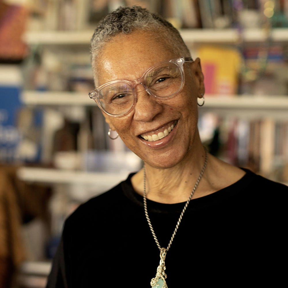 A multi-gendered, gray-haired, African-American lesbian artist poses in front of a bookshelf with a big smile. They are wearing a black shirt and a necklace with a light blue stone.
