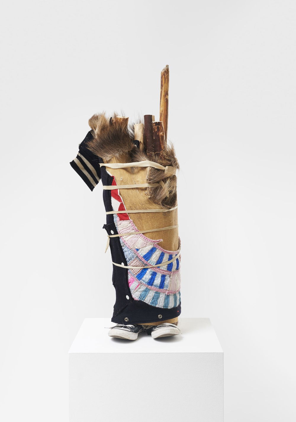 A sculpture vaguely shaped like a person sits atop a white pedestal. Converse sneakers peek out of the bottom of a trunk made of textiles and quilts wrapped with bands of material around a bundle of sticks. The sleeve of a T-shirt is visible at top left.