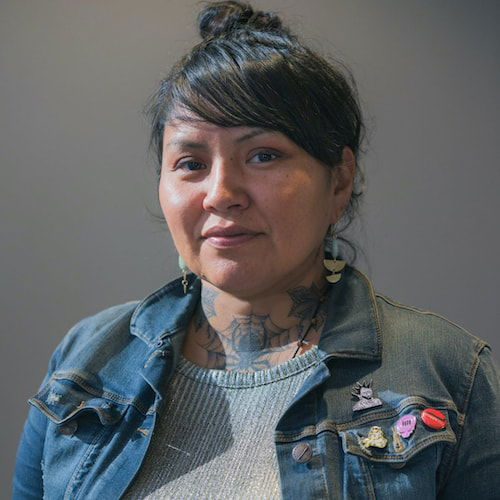 A headshot of a woman with her dark hair pulled into a bun on her head. She wears geometric earrings and a denim jacket adorned with enable pins on the pocket. She has a tattoo of a spider on her neck, its web visible over her shirt.