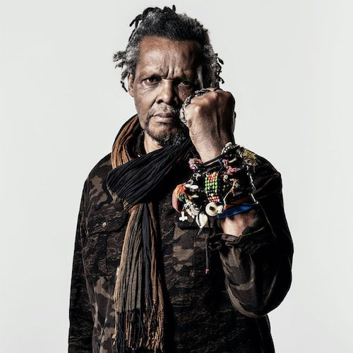 A portrait of a Black man standing in front of a white backdrop. He stares directly at the viewer with his left fist raised in front of his face, almost in a boxing position. However, instead of boxing gloves, his hand is adorned with silver rings and on his wrist he wears several bracelets made of plastic, beads, threads, and various fibers. On his face he wears a look of gentle concentration. His hair is greying on his head and goatee.