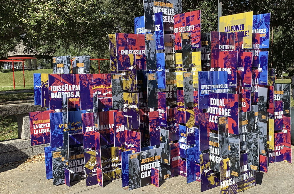 An outdoor space where dozens of medium-sized protest signs are stacked to create a mass of colorful cardboard resembling a jungle gym. Each individual poster has imagery printed in primary colors and white text with messages such as "Justice for Freddie Gray."