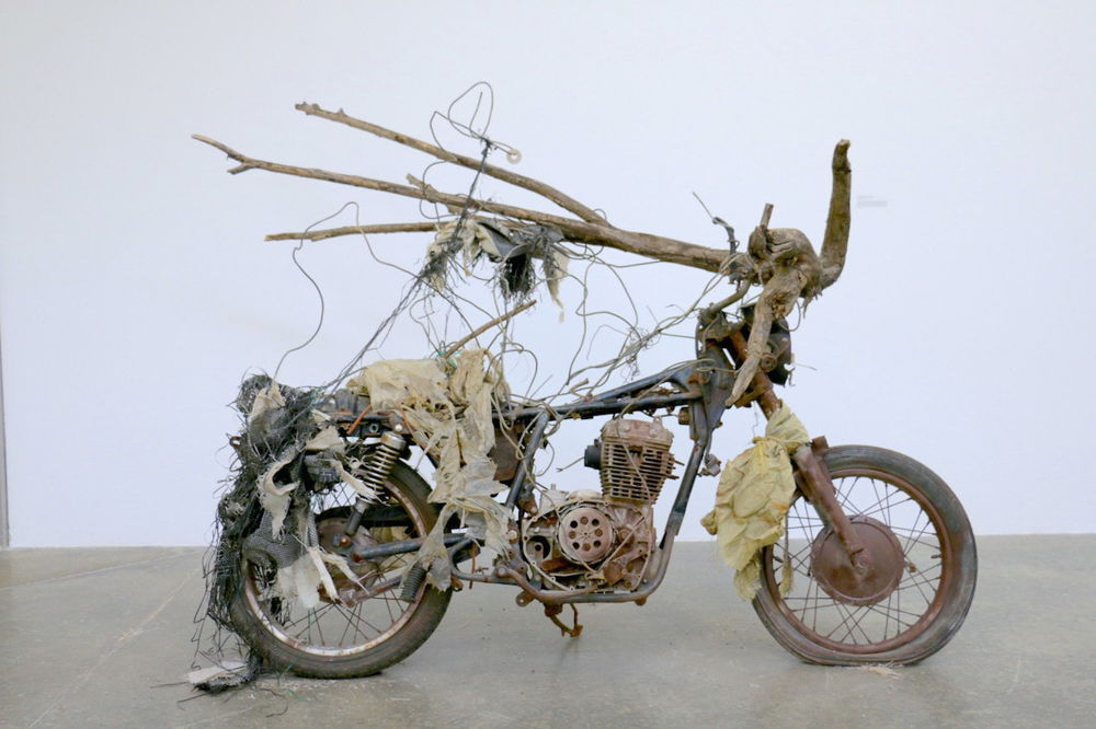 A photograph of an old rusted motorbike with a long branch of wood tied to its handles. Pieces of old plastic are tied to the front wheel and the seat; wires and threads weave in and out of the frame and the wood.