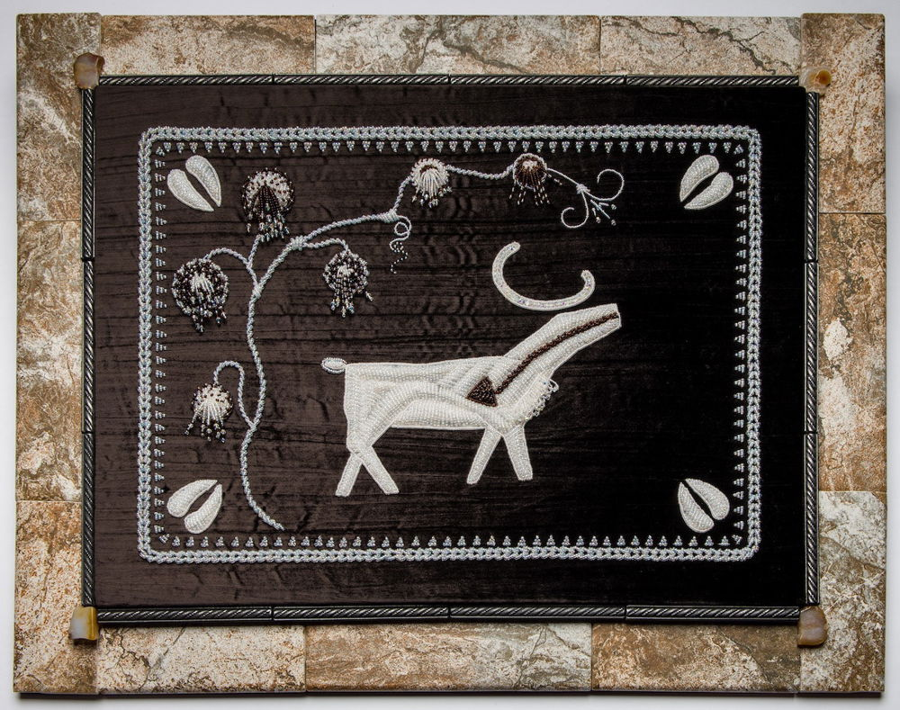 White and black beaded embroidery on a dark background. The image is a flat and stylized image of a caribou standing under a flowering vine. The embroidered scene is framed by a brown marbled background.