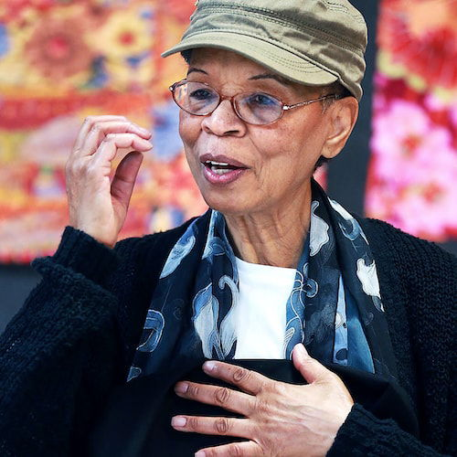 A candid portrait of a Black woman wearing a tan cap and glasses with colorful quilts out of focus behind her. She holds a quilt to her chest with one hand and gestures with the other as if in the middle of a conversation.