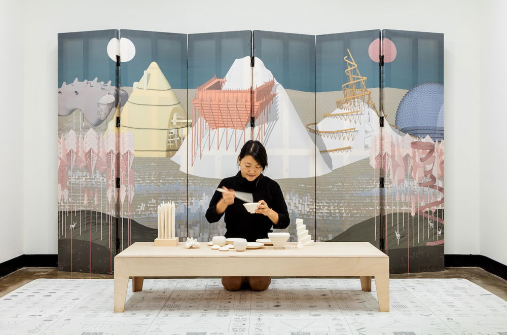 [ID: A woman kneels at a low table set with white ceramic dishes, holding a bowl and chopsticks. A folding screen is open behind her, depicting several mountains with various walls, ramps, and other constructions built onto them.]
