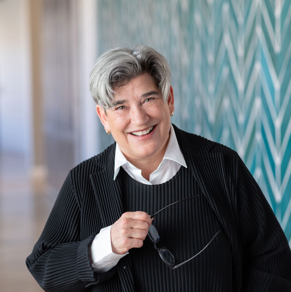 A white woman with short, salt-and-pepper hair smiles widely, holding a pair of glasses in one hand. She stands in front of a patterned wall.