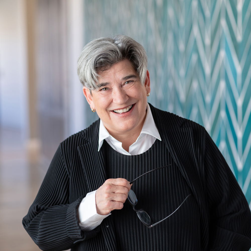 A white woman with short, salt-and-pepper hair smiles widely, holding a pair of glasses in one hand. She stands in front of a patterned wall.